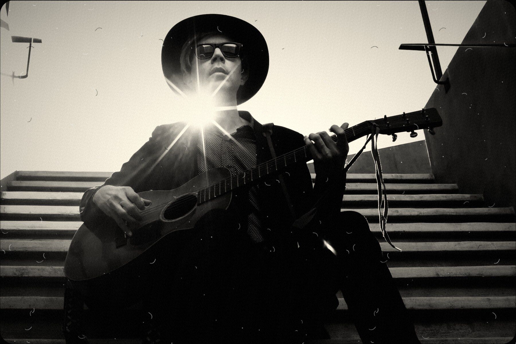 A guitarist in a wide-brimmed hat and sunglasses on a flight of steps, the sun bursting behind him